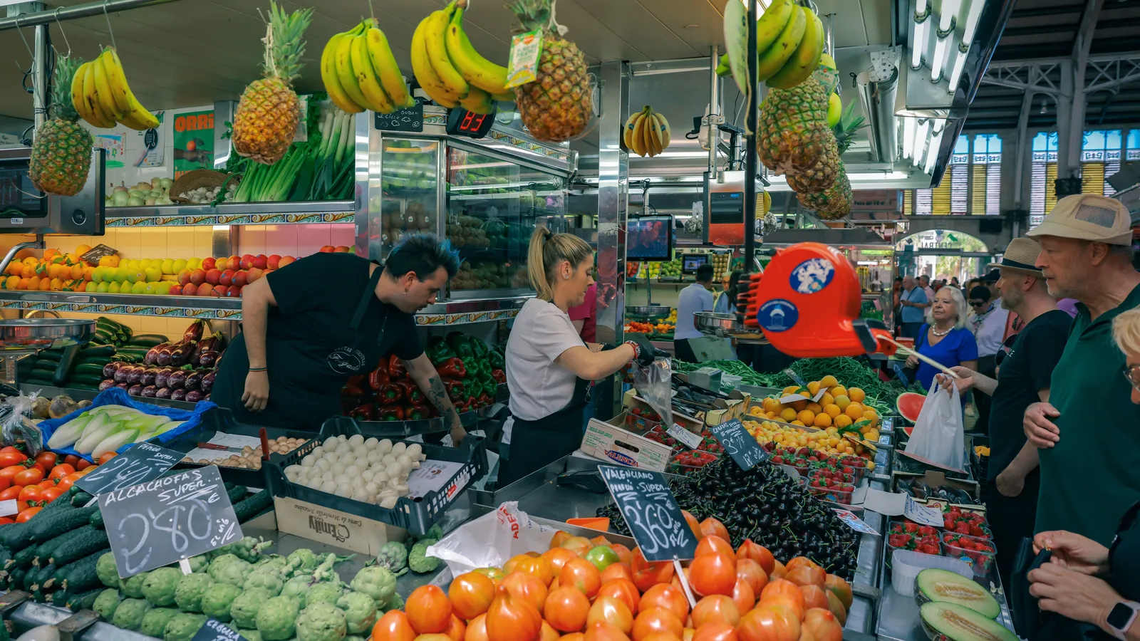 Vendors serving customers at a fruit and vegetable stall inside a covered food market in Spain