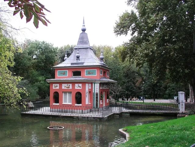 A small pink pavilion with a pointed roof and wooden balcony standing at the edge of a small pond surrounded by green trees