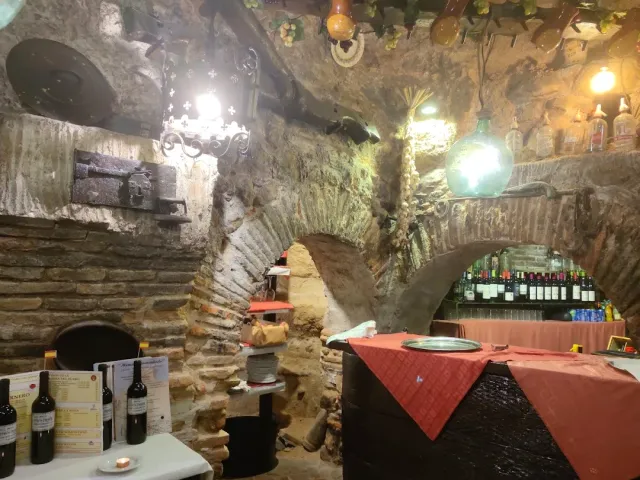 An arched stone cellar with wine bottles, hanging lamps and a table with a red cloth
