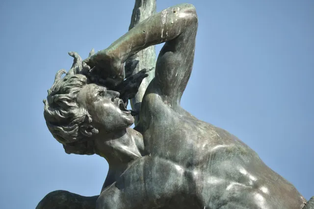 A bronze sculpture of a winged figure falling backwards, coiled by a serpent, against a background of treetops