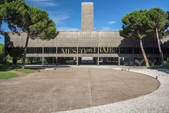 A dark modern building on pillars among pine trees, with a wide gravel forecourt in front