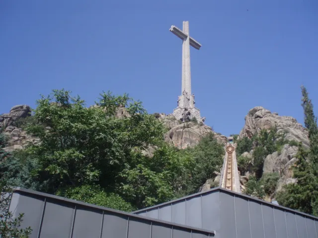A giant stone cross standing atop a rocky wooded hill, with a funicular line climbing towards it