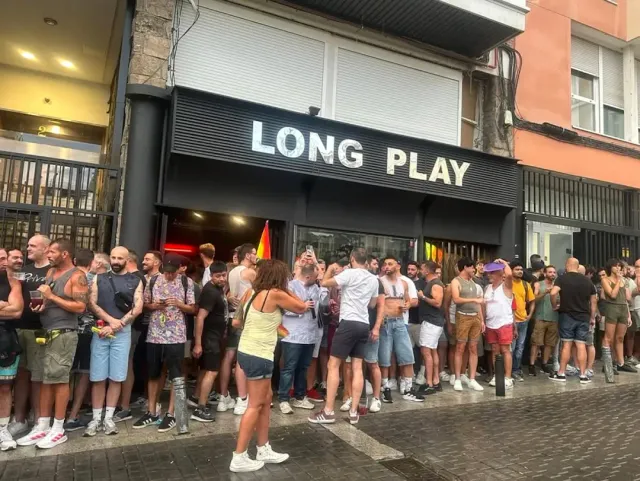 A queue of people on the sidewalk in front of a black frontage with a large letter sign