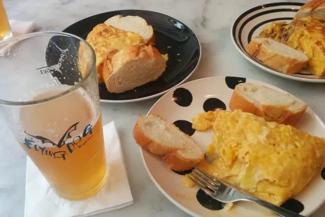 Plates of potato omelette with slices of bread, beside a glass of beer on a marble table