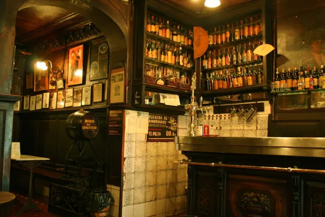 An old dark wooden bar with lit shelves of bottles, ceramic tiles and framed pictures on the walls
