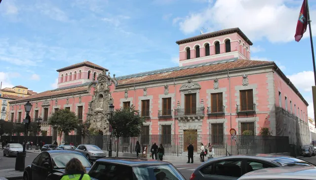 A long pink brick building with an ornate carved Baroque doorway behind an iron fence on a city street