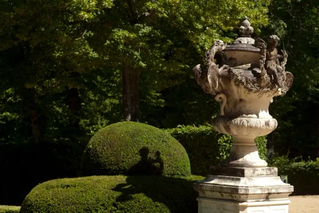 A carved stone urn on a tall pedestal at the end of a clipped hedge avenue, surrounded by dense green trees