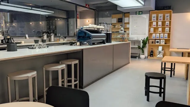 A long white counter with a blue espresso machine, stools and a shelf of coffee bags