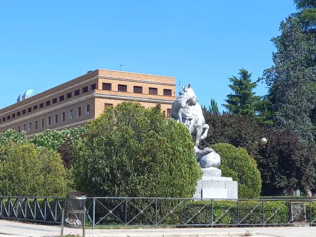 A stone sculpture of animals on a pedestal, behind a fence and shrubs, with a large brick building behind