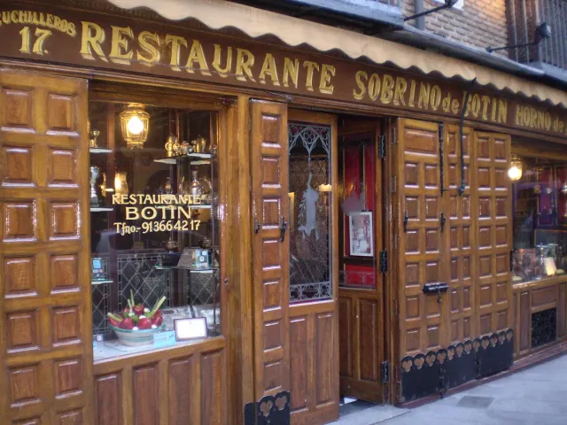 A dark wooden restaurant front with a gilded sign and a lit menu at the entrance on a stone street