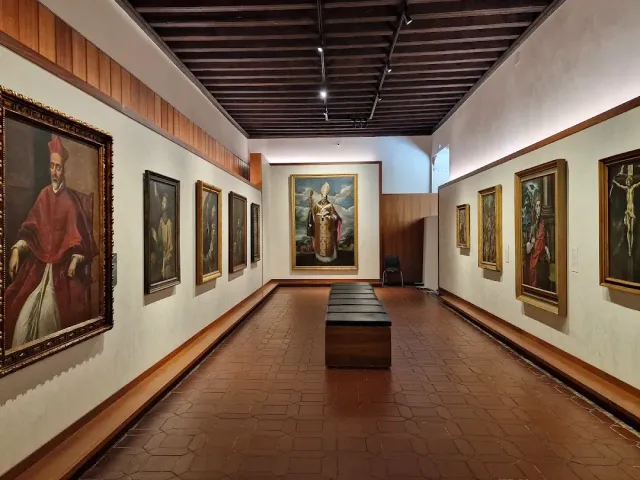 An exhibition hall with a wooden beamed ceiling, a terracotta tiled floor and portrait paintings on the walls