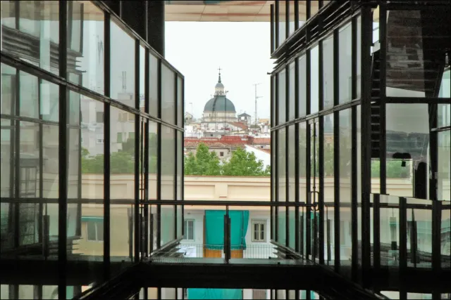 A modern glass and steel courtyard with red beams, an old building dome visible through the windows