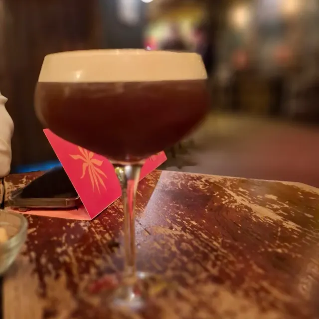 A dark cocktail glass with a layer of foam on a worn wooden table in dim light