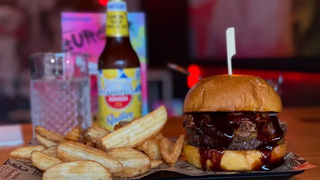 A burger in dark sauce beside thick-cut fries and a bottle of beer on the table