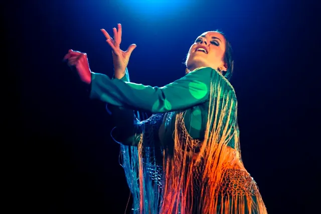 A dancer in a dress with long fringes spreading her arms on a dark stage
