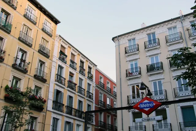 Yellow and white house facades with iron balconies and flower pots, and a red metro sign at the corner