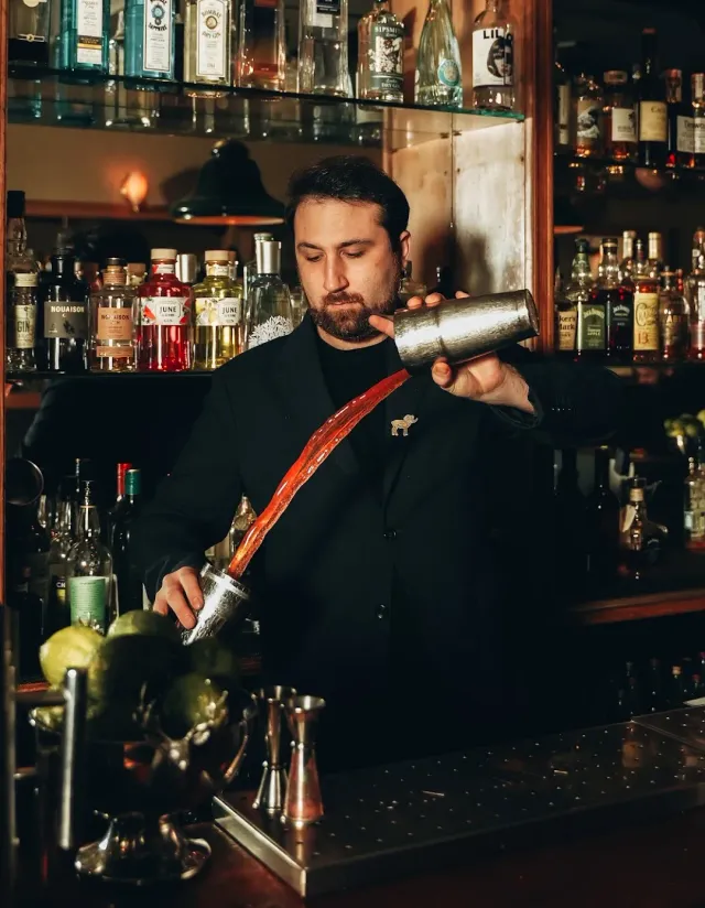A bartender in a black jacket pouring a drink from a shaker into a glass behind the counter