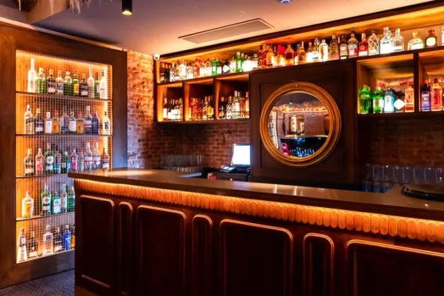 A wooden counter facing lit bottle shelves, a round mirror and a brick wall