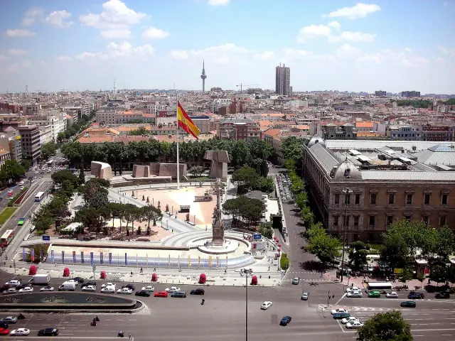 A view from above of a broad square with a giant flag, gardens and a city skyline behind