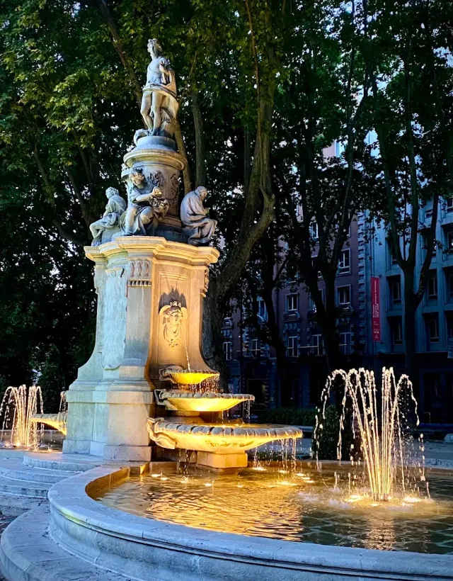 A lit stone fountain among trees at dusk, with a statue on top and jets of water