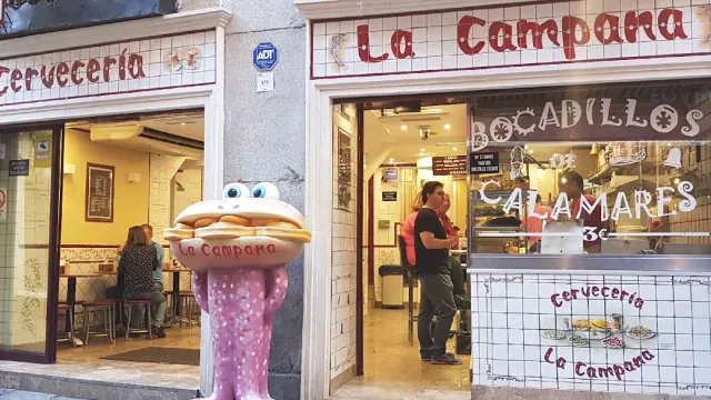 A bar front with tiled lettering, a sandwich figure at the entrance and a counter with a worker behind it