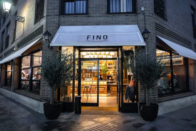 A brick facade with a white awning, potted olive trees and a lit bar seen through the windows in the evening