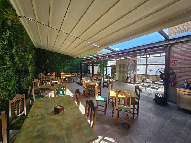 A patio covered by an awning with wooden tables, green chairs and a plant wall