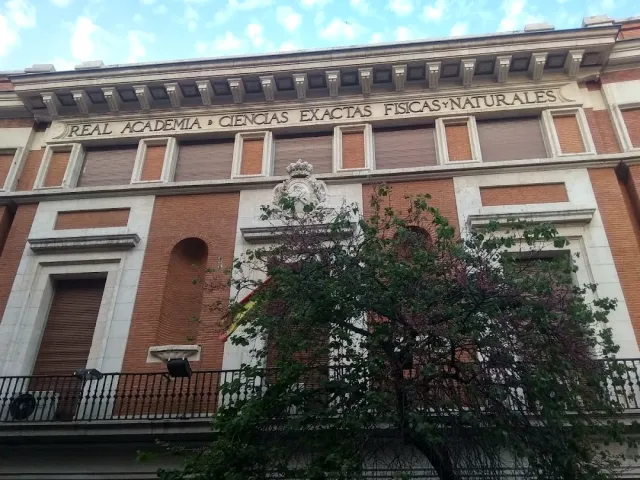 A red brick facade with an inscription carved in stone, a sculpted emblem and a tree in front