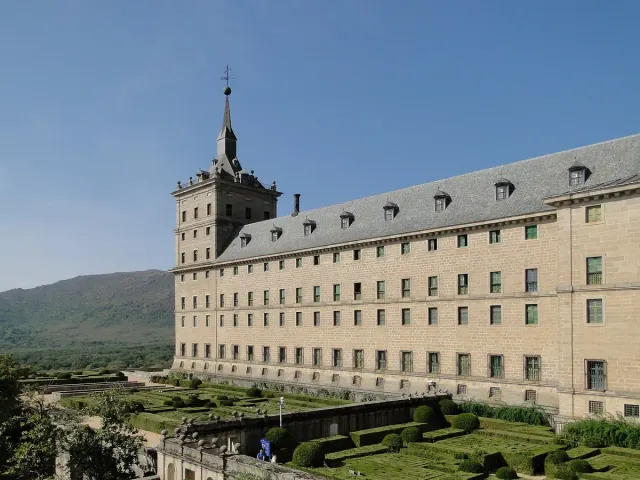 An immense grey stone complex with corner spires against green hills and mountains