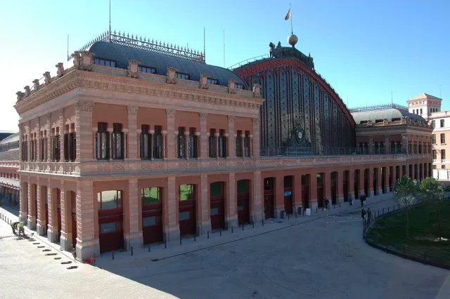 A red brick railway station facade with a clock tower and an iron and glass hall alongside