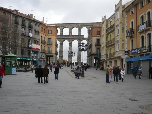 A paved square with pedestrians and colourful buildings, with tall aqueduct arches rising behind
