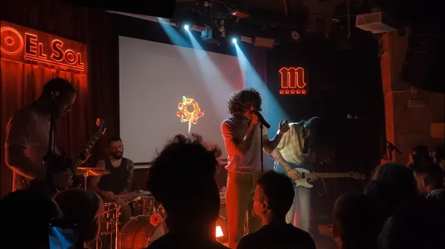 A small club stage lit in coloured lights with musicians and a silhouetted crowd