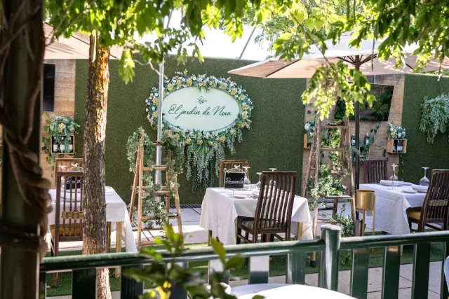 A garden terrace with a green hedge wall, tables in white cloths and parasols among the trees