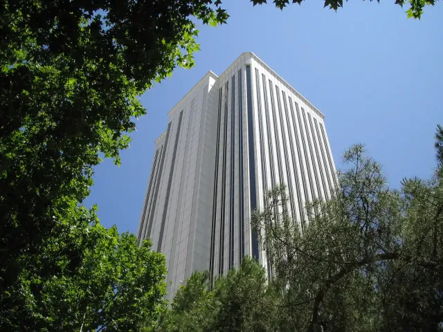 A white ribbed office tower rising above green treetops against a blue sky