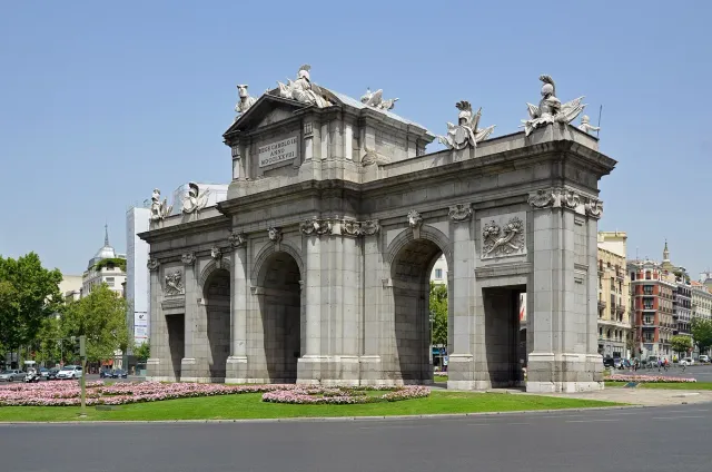 A neoclassical stone triumphal gate with five openings and sculptures on the pediment, ringed by pink flower beds