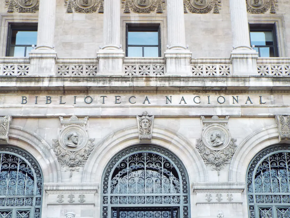 A pale stone facade with three arched entrances, carved portrait medallions and incised lettering above them