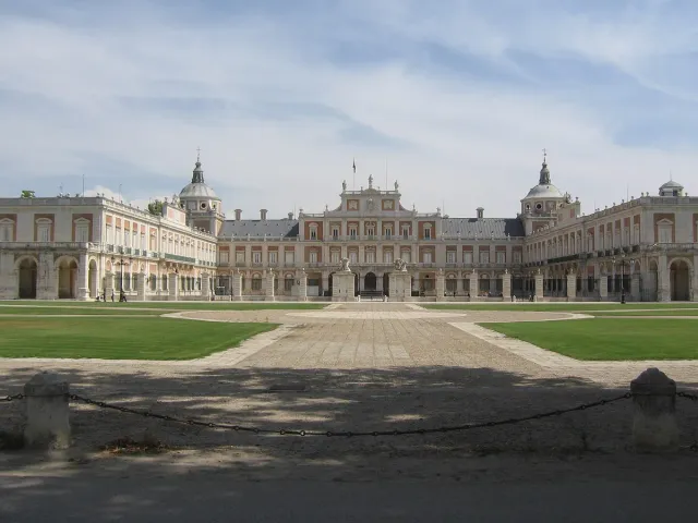 A pale symmetrical palace with a slate roof fronted by a wide forecourt and lawns