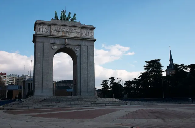 A large pale stone triumphal arch topped by a sculptural group, standing on an open plaza