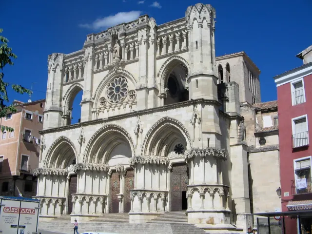 A Gothic cathedral facade in pale stone with arched windows, flanked by colourful houses