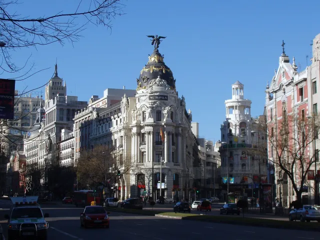 A corner building with a gilded dome topped by a winged statue on a busy city avenue