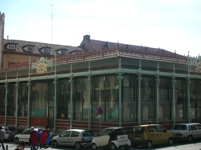 An iron and glass market hall with a green frame, surrounded by buildings and parked cars