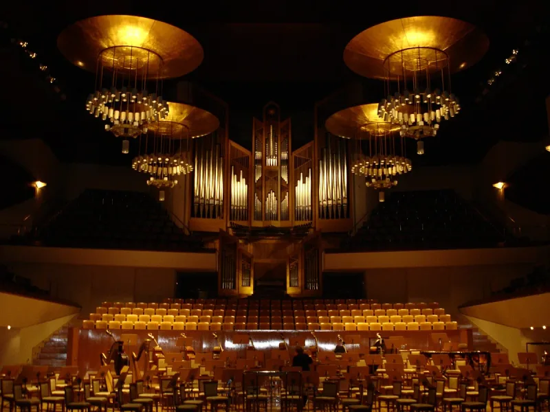 A concert hall with a large pipe organ, gold chandeliers and orchestra chairs set out on stage