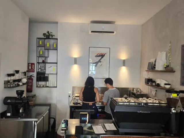 Two workers behind a narrow counter with an espresso machine and shelves on the wall