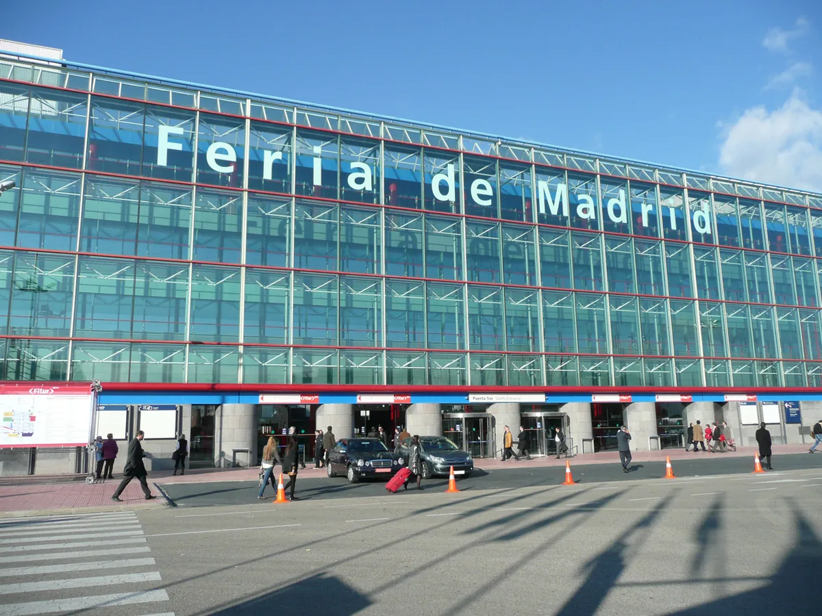 The long glass and steel front of an exhibition centre with large pale blue lettering under a clear sky