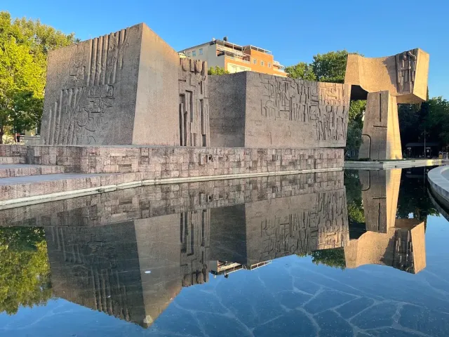 Large carved concrete blocks reflected in a shallow pool of water, among trees