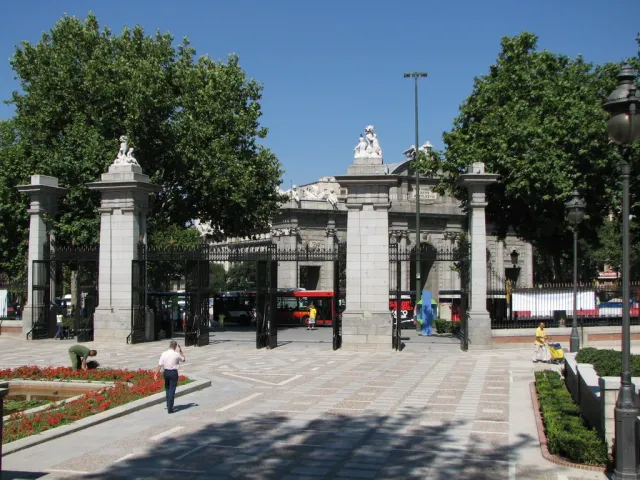 A monumental stone gate with columns and railings at a park entrance, facing a plaza and flower beds
