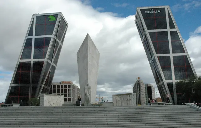 Two office towers leaning towards each other to form a gate above the road, with an obelisk between them