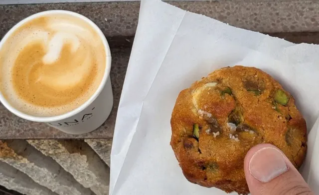 A paper cup of coffee with a pattern in the foam and a hand holding a cookie with pistachios