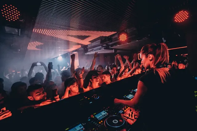 A DJ facing a dancing crowd with raised hands under red lights