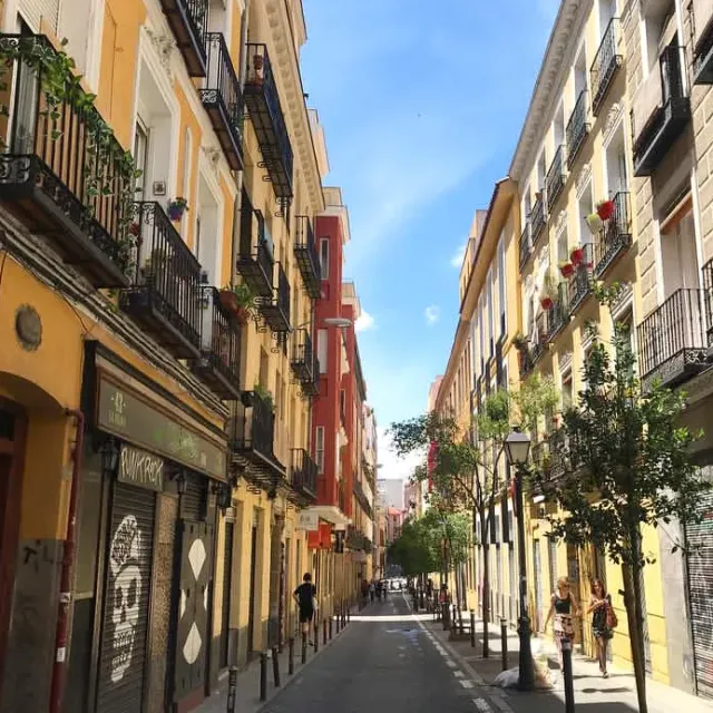 A narrow street with yellow and red facades, iron balconies with plants, and passers by